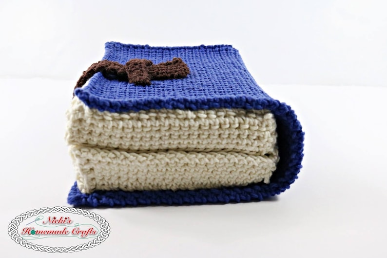 Pode incluir: Um travesseiro de croch&ecirc; em forma de livro com uma capa azul e um marcador de p&aacute;gina de croch&ecirc; marrom. O travesseiro &eacute; feito de duas camadas de tecido de croch&ecirc;, uma branca e uma azul.