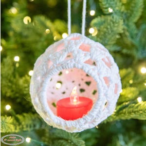 May include: White crocheted Christmas bauble with a red tea light candle inside. The bauble is hanging on a Christmas tree.