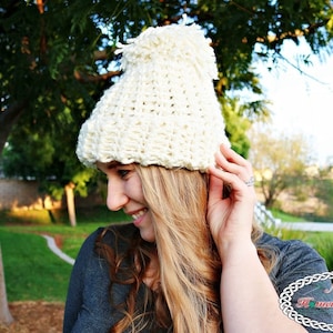 Op de afbeelding: Een witte gebreide beanie met een pompon bovenop. De beanie wordt gedragen door een persoon met lang blond haar. De persoon draagt een donkergrijs shirt.