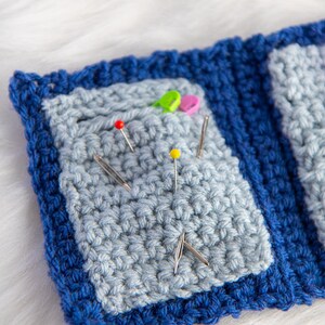 Pu&ograve; includere: Un cuscino per spilli in crochet blu con un centro blu chiaro e una variet&agrave; di spilli e aghi colorati.