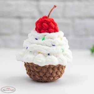 Könnte beinhalten: Handgehäkelter Cupcake mit brauner Basis und weißem Zuckerguss. Der Zuckerguss ist mit bunten Streuseln verziert und mit einer roten Kirsche gekrönt. Ein skurriles Dekorationsstück.