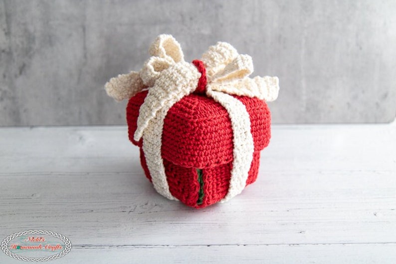 May include: A crocheted red gift box with a white crocheted bow. The box is sitting on a white wooden surface.