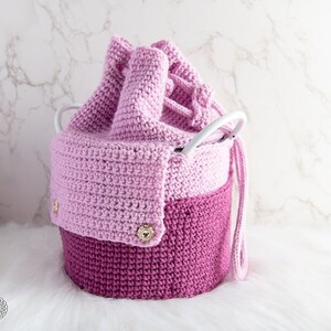 Puede incluir: Una cesta de almacenamiento de crochet rosa y morado con un asa blanca y botones dorados. La cesta est&aacute; hecha de dos tonos de rosa diferentes y tiene un cierre de cord&oacute;n.