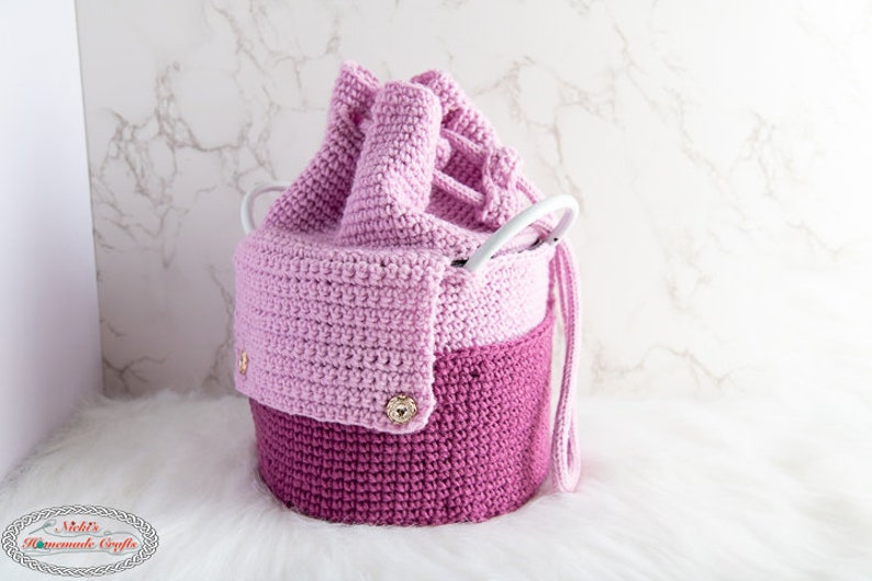 Puede incluir: Una cesta de almacenamiento de crochet rosa y morado con un asa blanca y botones dorados. La cesta est&aacute; hecha de dos tonos de rosa diferentes y tiene un cierre de cord&oacute;n.