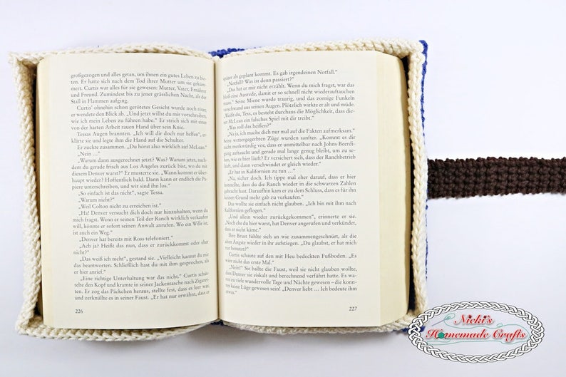 Pode incluir: Uma capa de livro de croch&ecirc; branca e azul com um marcador de p&aacute;gina de croch&ecirc; castanho. A capa do livro est&aacute; aberta numa p&aacute;gina com texto alem&atilde;o. O texto diz "Was ich dir noch sagen muss, ist das: Nie mehr kannst du es mir verzeihen." A capa do livro &eacute; um artesanato feito &agrave; m&atilde;o.
