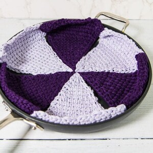 Könnte beinhalten: Ein lila und lavendelfarbener, gestrickter Topflappen in Form einer Blume, der in einer schwarzen Bratpfanne liegt.