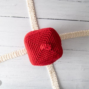 May include: A red crocheted hat with a small pom-pom on top. The hat is lying on a white wooden surface with two white crocheted strips.