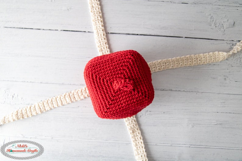 May include: A red crocheted hat with a small pom-pom on top. The hat is lying on a white wooden surface with two white crocheted strips.