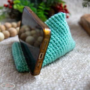 Puede incluir: Un soporte para teléfono de color azul turquesa tejido a crochet con un teléfono inteligente apoyado en él. El soporte está hecho de hilo y tiene forma triangular.