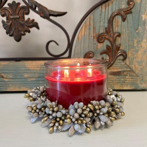 May include: A red candle in a glass jar sits on a decorative silver and gold wreath candle ring.