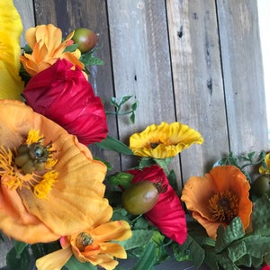 Puede incluir: Una guirnalda floral con flores rojas, naranjas y amarillas, hojas verdes y peque&ntilde;as vainas de semillas marrones. La guirnalda est&aacute; drapeada sobre una superficie de madera desgastada.