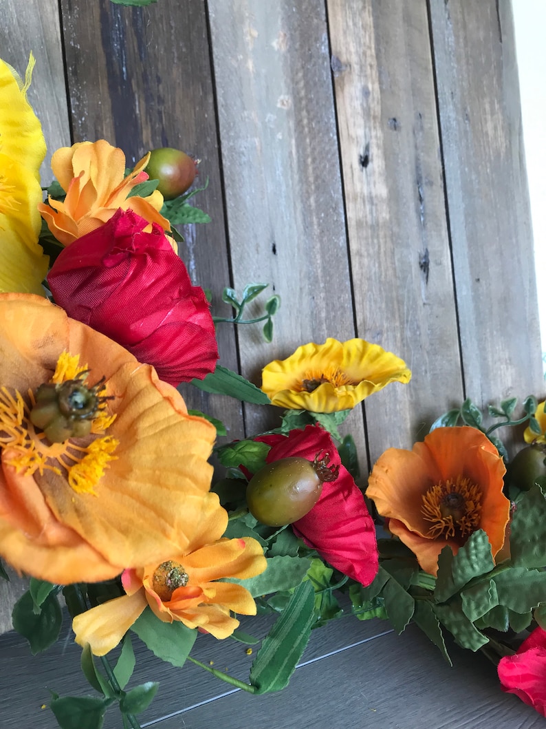 Puede incluir: Una guirnalda floral con flores rojas, naranjas y amarillas, hojas verdes y peque&ntilde;as vainas de semillas marrones. La guirnalda est&aacute; drapeada sobre una superficie de madera desgastada.