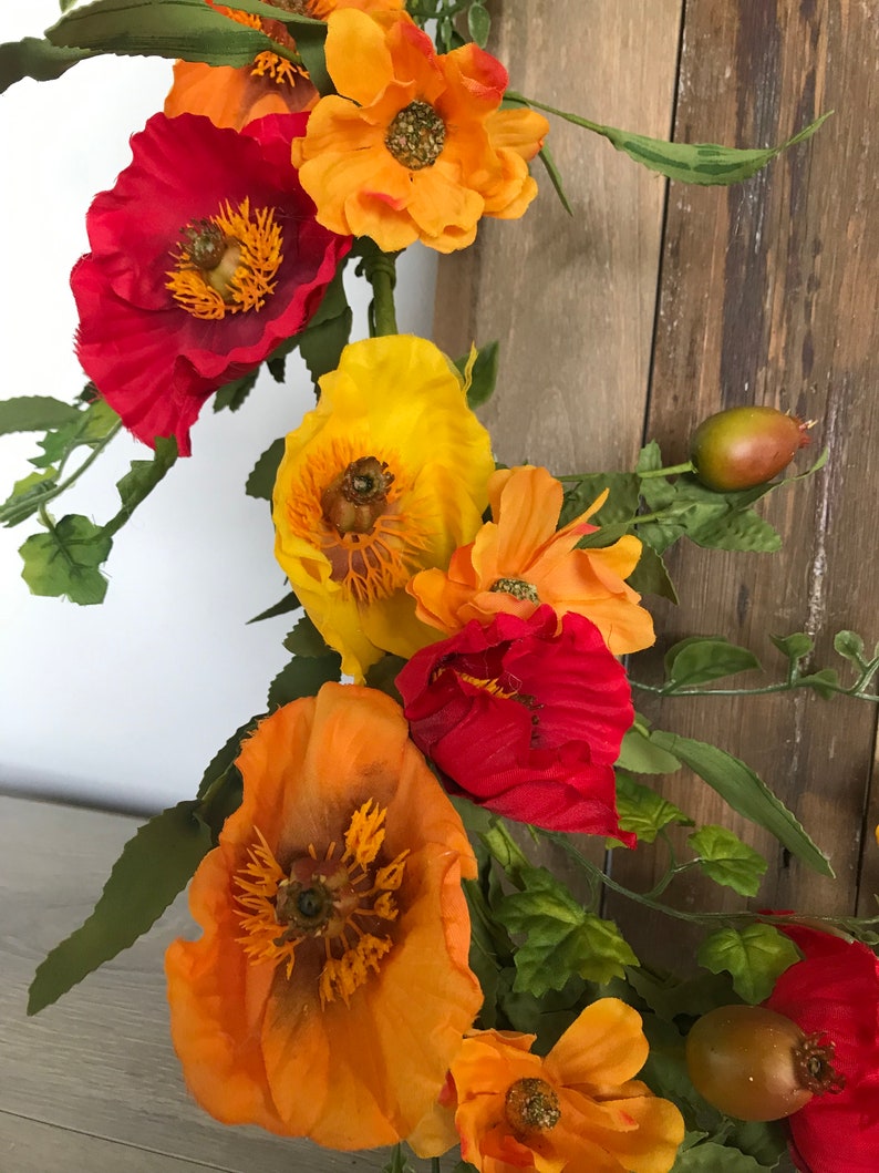 Puede incluir: Una corona floral con flores de amapola rojas, naranjas y amarillas y hojas verdes.
