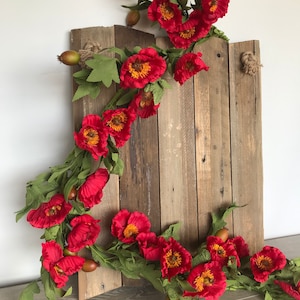 May include: A rustic wooden sign adorned with a vibrant garland of red poppies with yellow centers and green leaves. The sign is made of weathered wood planks, with a natural rope detail at the top. The floral arrangement adds a touch of color.