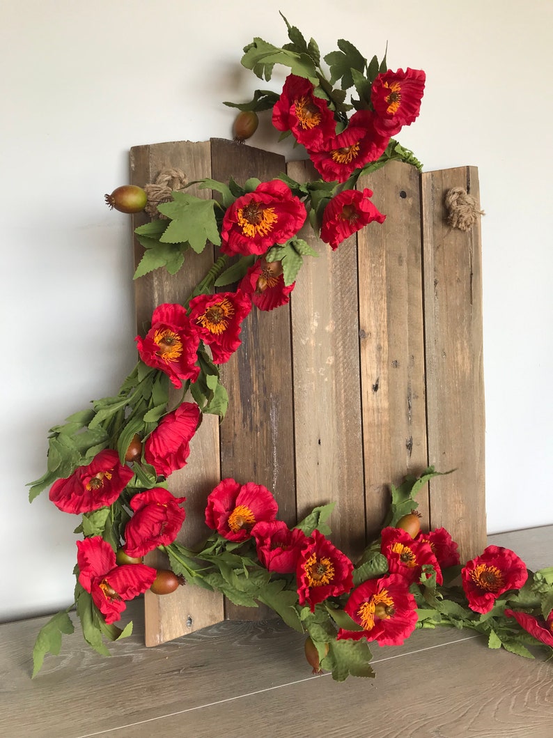 May include: A rustic wooden sign adorned with a vibrant garland of red poppies with yellow centers and green leaves. The sign is made of weathered wood planks, with a natural rope detail at the top. The floral arrangement adds a touch of color.