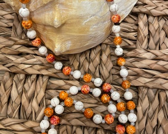 May include: A long necklace made of white and orange beads with brown accents. The beads are round and have a speckled pattern. The necklace is draped over a seashell.