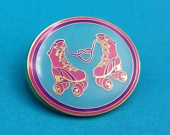 Könnte beinhalten: Eine goldfarbene Emaille-Anstecknadel mit zwei rosa Rollschuhen, die mit einem rosa Band zusammengebunden sind. Die Schuhe befinden sich auf einem hellblauen Hintergrund mit einem violetten und rosa Rand.
