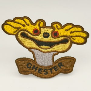May include: Embroidered patch featuring a cartoon character with yellow antlers, red eyes, and a smiling mouth. The patch has a brown border and the name "CHESTER" embroidered on a banner below the character.