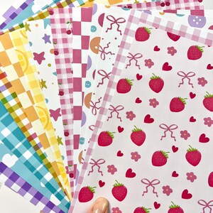 May include: A collection of patterned paper sheets in various designs. Patterns include gingham, strawberries, bows, cherries, and lemons. Colors include pink, yellow, blue, and purple. The sheets are fanned out, showcasing the variety of designs.