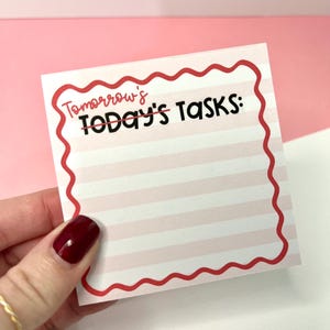May include: A square notepad sheet with light pink horizontal stripes and a red wavy border. The text reads Tomorrow's with a line through it, followed by Today's Tasks: in black ink. The paper has a white background with faint lines.