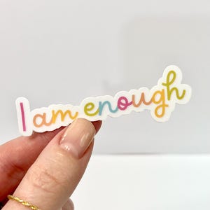Può includere: Un adesivo bianco con la scritta "I am enough" in un carattere colorato e scritto a mano. Il testo passa dal rosa al giallo, al verde e al blu. L'adesivo è mostrato su uno sfondo bianco.