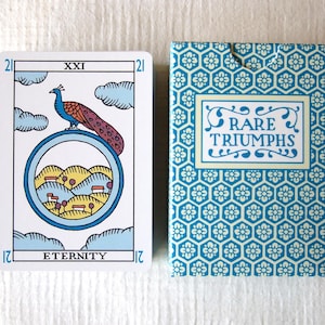 May include: A tarot card labeled XXI ETERNITY featuring a peacock perched on a circular frame enclosing a landscape with hills and houses, alongside a blue and white patterned tuck box with the text RARE TRIUMPHS printed on the front panel.