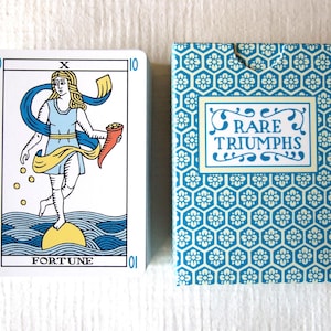 May include: A deck of tarot cards featuring a card labelled FORTUNE with a figure standing on a yellow sphere above blue waves, holding a cornucopia. Beside it is a blue and white box with a hexagonal floral pattern and the text RARE TRIUMPHS.