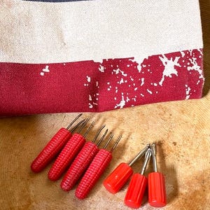 May include: A patriotic-themed kitchen towel with a blue top section featuring white stars, a white middle section, and a red bottom section. Six red corn cob holders are also visible.