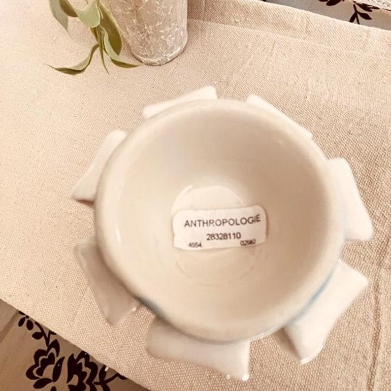 Puede incluir: Un cuenco de cer&aacute;mica blanco con un dise&ntilde;o floral. El cuenco tiene forma circular con extensiones en forma de p&eacute;talo alrededor del borde. El texto "ANTHROPOLOGIE" es visible en una etiqueta dentro del cuenco.