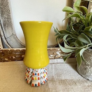 Puede incluir: Un jarr&oacute;n amarillo brillante con una parte superior acampanada se asienta sobre una base decorativa. La base es blanca con detalles en rojo, azul y amarillo. El jarr&oacute;n es una pieza de acento vibrante.