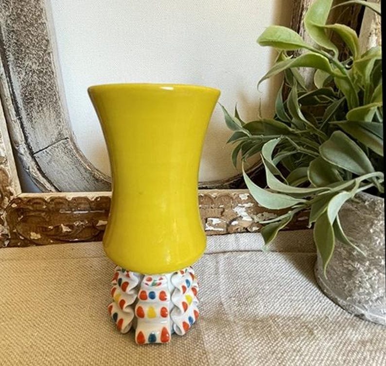 Puede incluir: Un jarr&oacute;n amarillo brillante con una parte superior acampanada se asienta sobre una base decorativa. La base es blanca con detalles en rojo, azul y amarillo. El jarr&oacute;n es una pieza de acento vibrante.