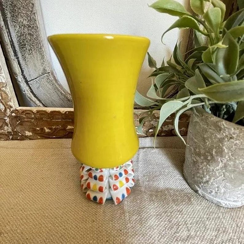 Puede incluir: Un jarr&oacute;n de cer&aacute;mica amarillo brillante con la parte superior acampanada y una base decorativa. La base est&aacute; adornada con un volante de tela blanca con lunares y tri&aacute;ngulos de colores. El jarr&oacute;n se exhibe sobre una superficie de tono neutro.