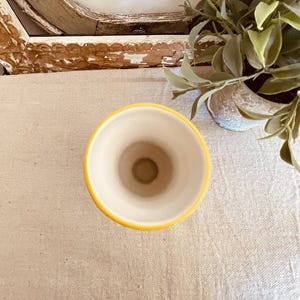Puede incluir: Un peque&ntilde;o jarr&oacute;n de cer&aacute;mica con borde amarillo e interior crema. El jarr&oacute;n tiene un dise&ntilde;o sencillo y probablemente se utiliza para exhibir flores u otros objetos decorativos. El jarr&oacute;n est&aacute; sobre una superficie de color neutro.