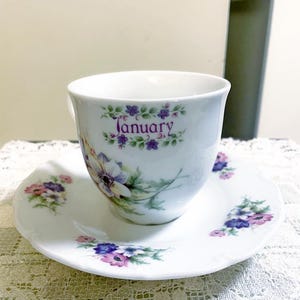 May include: A white porcelain teacup and saucer set featuring a delicate floral pattern with purple, blue, and pink flowers. The word January is printed in purple script on the front of the cup, surrounded by small green leaves and purple blossoms.