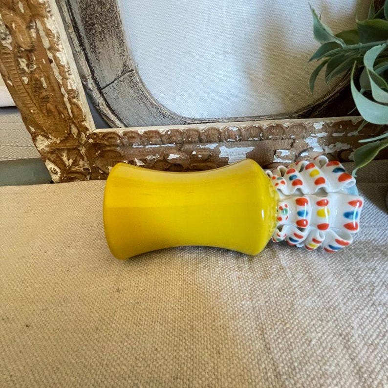 Puede incluir: Un objeto de vidrio amarillo brillante con forma de reloj de arena curvado. Junto a &eacute;l, un peque&ntilde;o recipiente de cer&aacute;mica blanca con un dise&ntilde;o a rayas de colores rojo, azul y amarillo. Los objetos est&aacute;n sobre una superficie de color neutro.