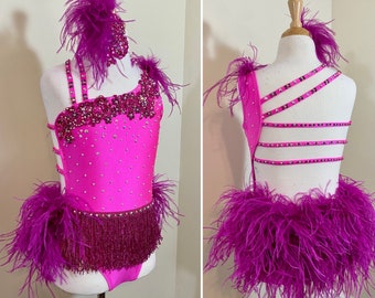 May include: A vibrant fuchsia dance costume with a one-shoulder design. The bodice is embellished with rhinestones and a fringe hem. The costume features a feathered skirt and matching headpiece, displayed on a mannequin.