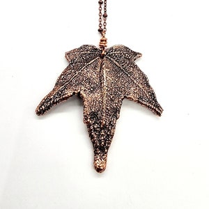 May include: A copper necklace with a detailed leaf pendant. The leaf is textured and has a pointed tip. The chain is made of small copper beads.