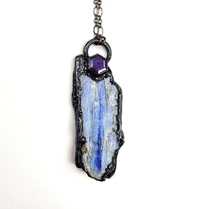 May include: A handcrafted pendant necklace featuring a blue kyanite stone with a dark, textured border. A small, purple gemstone is set above the kyanite, and the pendant hangs from a silver-colored chain. The necklace is displayed against a white background.