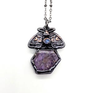May include: A dark metal pendant necklace featuring a moth-shaped top with copper-colored crescent moon accents and a small blue stone, connected to a large, irregular, faceted purple gemstone base held in a dark, textured metal setting.