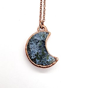 May include: A crescent moon pendant necklace. The pendant features a copper-colored frame and a blue and green stone. The necklace chain is also copper-colored. The necklace is displayed against a white background.