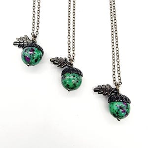 May include: Three necklaces with acorn pendants. Each pendant features a green and red speckled stone, a dark metal acorn cap, and a leaf detail. The chains are silver-colored and hang against a white background.