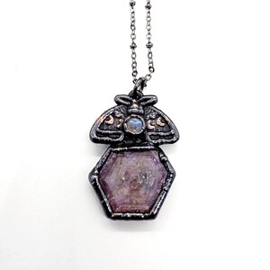 May include: A necklace featuring a dark metallic pendant shaped like a moth with a small iridescent round stone at its center, positioned above a larger hexagonal purple gemstone, all suspended from a thin silver-toned metal chain.