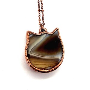 May include: A copper wire wrapped pendant necklace featuring a brown and white agate stone in the shape of a cat's head.