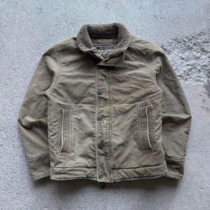 May include: Olive green button-up jacket with a brown, fuzzy collar. The jacket has two front pockets and a button closure. The brand name "Abercrombie & Fitch" is visible on a tag near the collar. The jacket is made of a durable material.