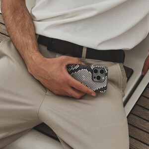 May include: A man wearing a white t-shirt and beige pants sits on a boat. He is holding a smartphone with a patterned case in his hand.
