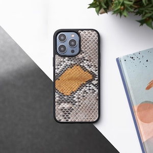 Puede incluir: Una funda de teléfono negra con un patrón de piel de serpiente en tonos marrones, grises y blancos. La funda tiene una protuberancia de cámara elevada con tres lentes.