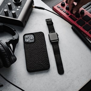 Könnte beinhalten: Ein schwarzes Leder-Handy-Etui mit strukturierter Oberfläche, eine schwarze Apple Watch mit schwarzem Armband, ein rotes MIDI-Keyboard mit schwarzen Tasten und Knöpfen, ein schwarzes Audio-Interface mit Knöpfen und Reglern, ein schwarzer Kopfhörer und eine braune Akustikgitarre mit schwarzem Schlagbrett.