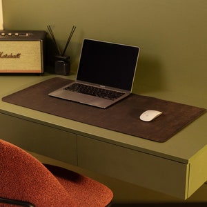 May include: A green desk with two drawers and a black metal frame. A laptop computer, a wireless mouse, and a small speaker are on the desk. A brown leather desk mat is on the desk. A red chair is in front of the desk.