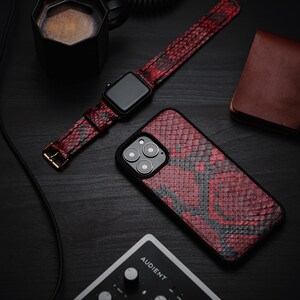 以下が含まれることがあります： 黒のApple Watchに、赤と黒のヘビ柄の腕時計バンドが付けられています。腕時計バンドは、黒いコーヒーカップ、茶色の革財布、黒いオーディオミキサー（「AUDIENT」と印刷されている）のある黒い表面の上に置かれています。