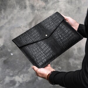 May include: A black leather clutch with a crocodile embossed pattern. The clutch has a flap closure with a silver button.